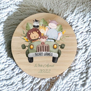 Puede incluir: Una placa redonda de madera con un jeep verde que lleva un león, un elefante, una jirafa, un hipopótamo, un mono y una cebra. El jeep tiene el nombre "Noah James" y la fecha "13 de abril de 2022" escrita en él.