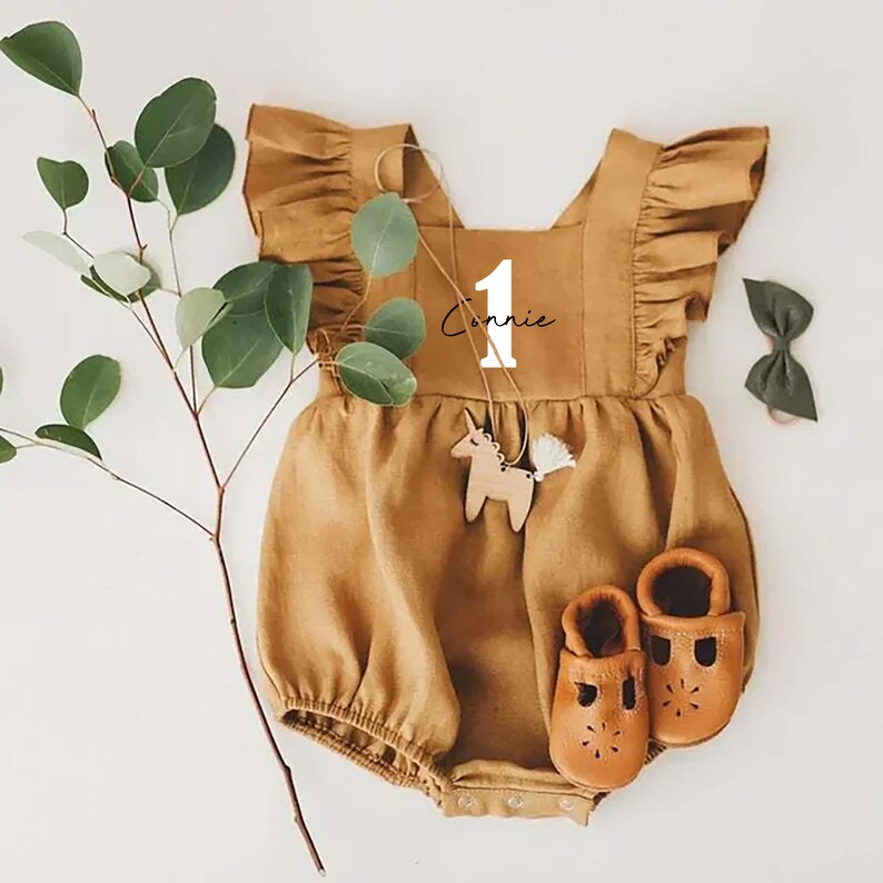 May include: A brown baby onesie with a white ruffle trim and the number "1" and the name "Connie" embroidered on the front. The onesie is paired with brown leather shoes and a wooden unicorn toy.