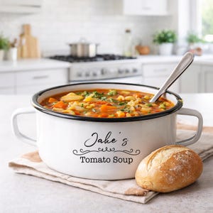 Può includere: Una zuppiera in smalto bianco piena di zuppa di pomodoro, un cucchiaio e un panino. La pentola ha finiture e manici neri, con la scritta "Jake's Tomato Soup". La zuppa contiene verdure ed erbe.