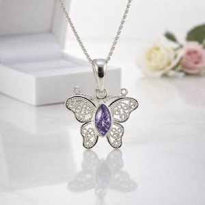 May include: A silver butterfly pendant necklace with a purple central stone. The butterfly has detailed filigree wings and hangs from a fine chain. The necklace is displayed on a white surface, with a white box and flowers in the background.