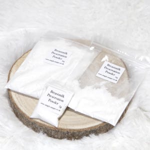 以下が含まれることがあります： 木のスライスの上に、「Breastmilk Preservation Powder」とラベルされた白い粉末が入った3つの透明なビニール袋。 袋には5g、10g、50gの粉末が入っています。 背景は柔らかく白いテクスチャのある表面です。