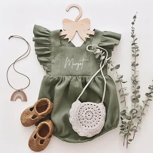 May include: A green linen baby romper with a ruffled neckline and a white embroidered name "Margot". The romper is hanging on a wooden hanger with a bow. A small crocheted bag and a pair of brown leather shoes are also pictured.