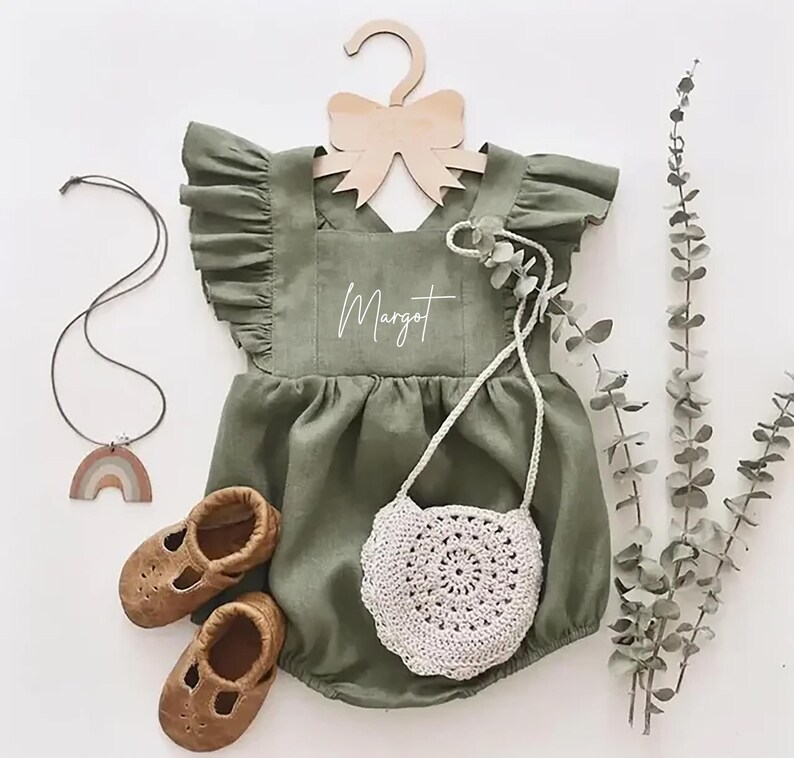 May include: A green linen baby romper with a ruffled neckline and a white embroidered name "Margot". The romper is hanging on a wooden hanger with a bow. A small crocheted bag and a pair of brown leather shoes are also pictured.
