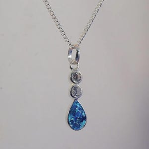 May include: A silver necklace with a pendant featuring three clear, round stones and a teardrop-shaped blue stone. The pendant hangs from a delicate chain, creating an elegant and eye-catching design.