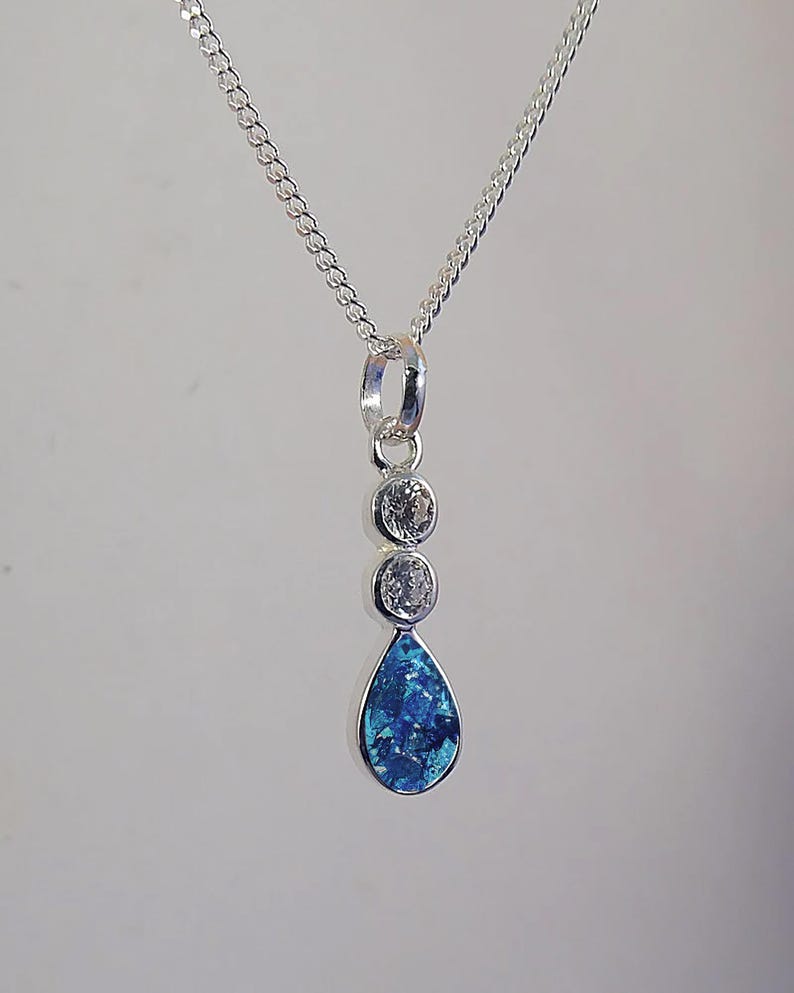 May include: A silver necklace with a pendant featuring three clear, round stones and a teardrop-shaped blue stone. The pendant hangs from a delicate chain, creating an elegant and eye-catching design.