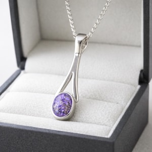 May include: A silver pendant necklace featuring a purple and gold oval gemstone. The pendant hangs from a fine chain and is displayed in a black jewellery box with a white lining.