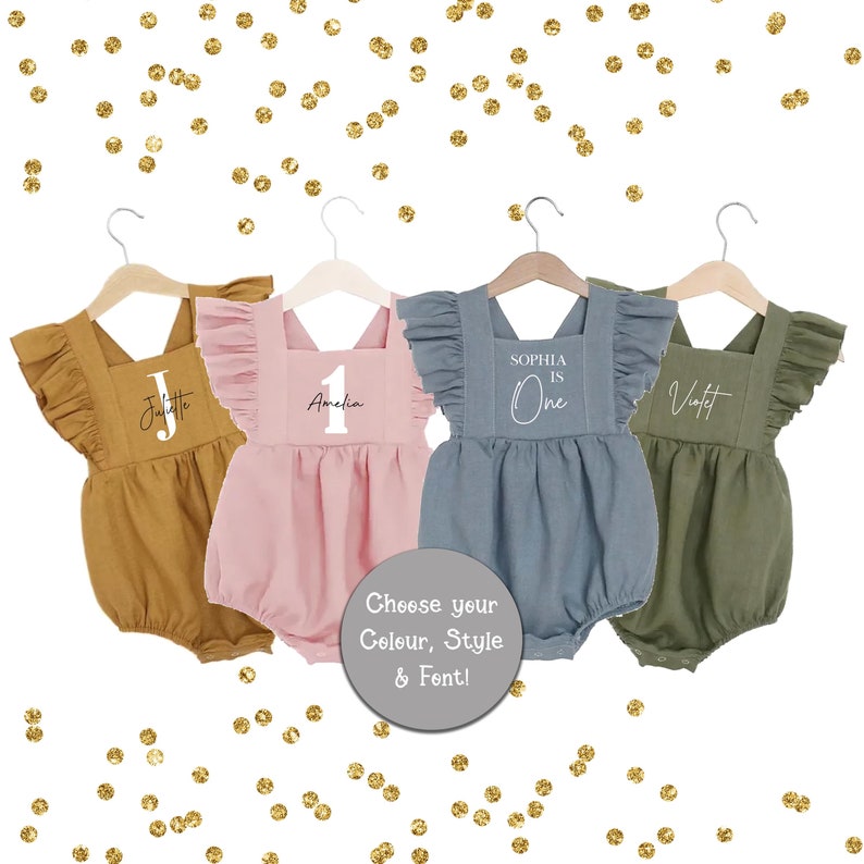 May include: Four personalized baby onesies in different colors, with the names Juliette, Amelia, Sophia, and Violet. The onesies have ruffles on the shoulders and are hanging on hangers. The text on the onesies says "Sophia is One", "Amelia 1", "Juliette", and "Violet".
