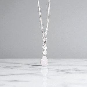 May include: A silver necklace with a pendant featuring two round, clear stones above a pear-shaped, light pink stone. The chain is a delicate curb link style, and the pendant hangs from a small loop.