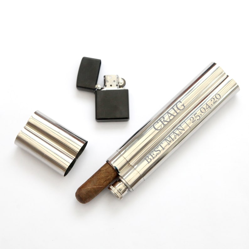 May include: A silver cigar tube with a brown cigar inside. The tube has an inscription that reads "CRAIG BEST MAN 25.04.20". A black lighter is also pictured.
