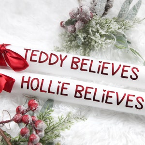 May include: Two white cardboard tubes with red lettering. The text on the tubes reads "Teddy Believes" and "Hollie Believes". The tubes are decorated with red ribbon bows. The tubes are on a white fluffy surface with a sprig of red berries and green leaves.