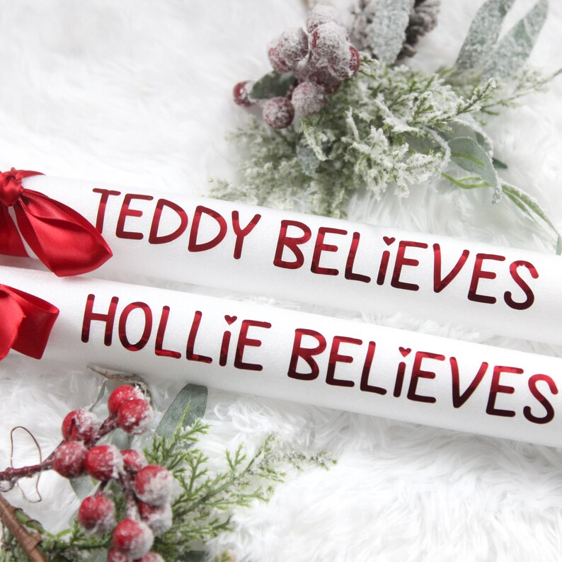 May include: Two white cardboard tubes with red lettering. The text on the tubes reads "Teddy Believes" and "Hollie Believes". The tubes are decorated with red ribbon bows. The tubes are on a white fluffy surface with a sprig of red berries and green leaves.