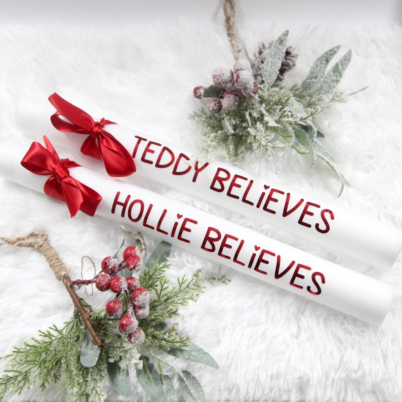May include: Two white rolled up paper tubes with red ribbon bows and the text "Teddy Believes" and "Hollie Believes" in red lettering. The tubes are on a white fluffy surface with two sprigs of greenery with red berries.