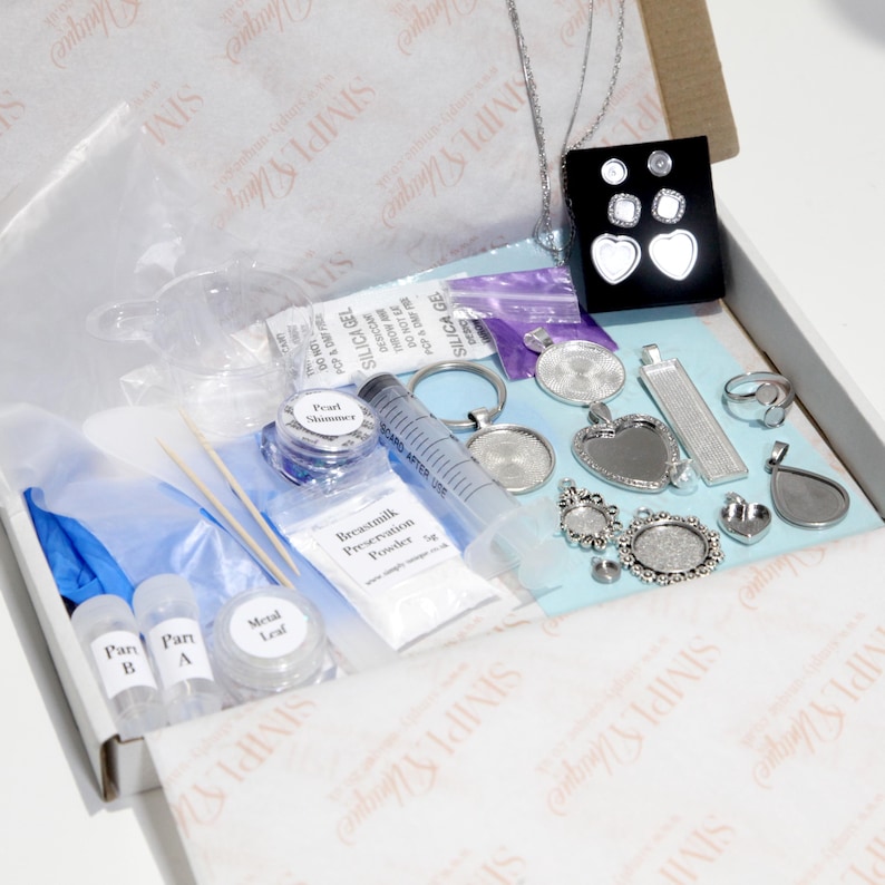 May include: A jewelry-making kit with various components, including pendants, earrings, a ring, and a syringe. The kit also contains small containers of powder, glitter, and other materials, all neatly arranged inside a cardboard box.