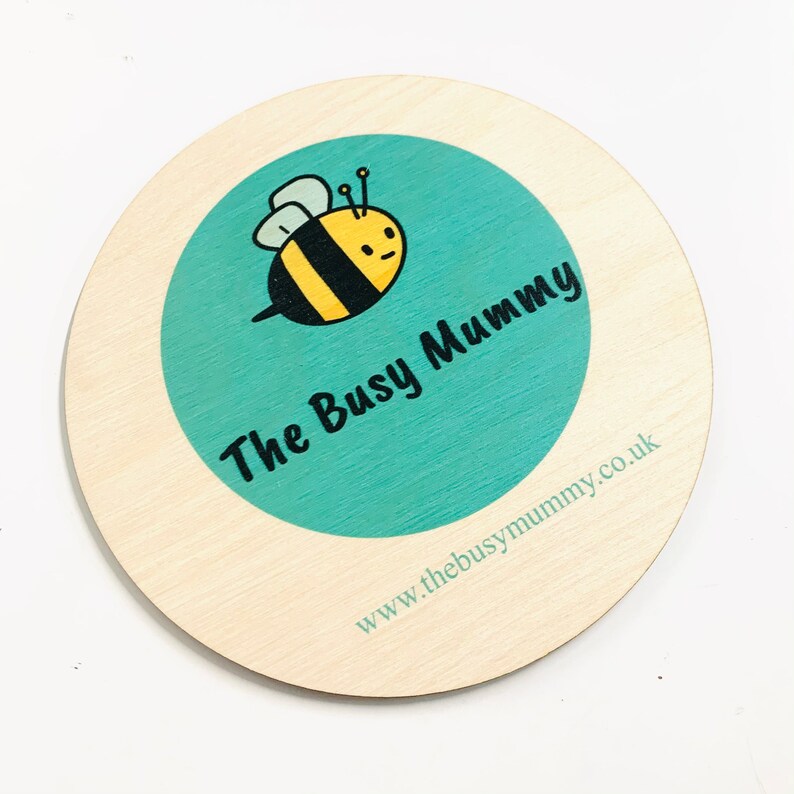 May include: A round wooden coaster with a turquoise circle in the center. The circle features a cartoon bee with black and yellow stripes and the text "The Busy Mummy". The bottom of the coaster has the text "www.thebusymummy.co.uk".