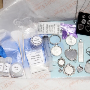 May include: A craft kit containing various jewelry-making supplies. Includes pendants, rings, earrings, a syringe, vials, and powders. The kit is packaged in a cardboard box with a clear plastic bag and blue gloves.