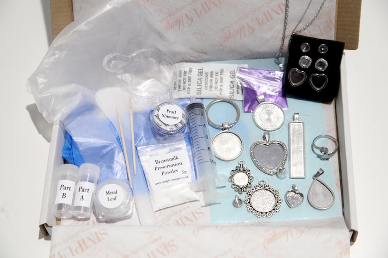 May include: A craft kit containing various jewelry-making supplies. Includes pendants, rings, earrings, a syringe, vials, and powders. The kit is packaged in a cardboard box with a clear plastic bag and blue gloves.
