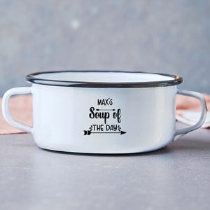 Pode incluir: Tigela de sopa esmaltada branca com acabamento preto e duas alças. A tigela tem o texto "MAX'S Soup of THE DAY" em preto e uma seta decorativa. A tigela está em uma superfície cinza.