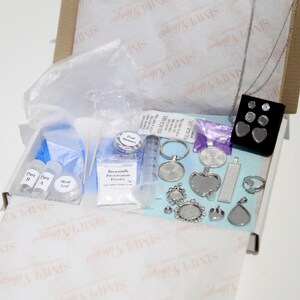 May include: An open cardboard box filled with jewelry-making supplies. The kit includes various silver-toned pendants, rings, and bezels, along with small bottles, a syringe, and packets of materials like pearl shimmer and metal leaf.