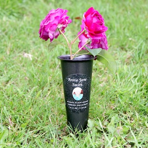 Puede incluir: Un jarrón de plástico negro con una pegatina personalizada que dice "Annie Jane Smith" y "Siempre en nuestros corazones para siempre y siempre 18.11.39 - 11.07.20". El jarrón está lleno de rosas rosas y está sentado en la hierba verde.