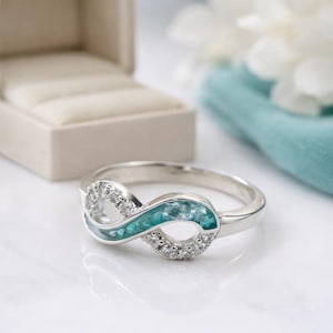Puede incluir: Anillo plateado con forma de infinito, con un centro turquesa y pequeñas piedras claras. El anillo se muestra sobre una superficie blanca, con una caja de anillos en el fondo.