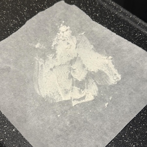 May include: A square of parchment paper with a white, textured substance spread across it. The paper sits on a dark, speckled surface, possibly a baking sheet or tray. The substance appears to be a paste or powder.