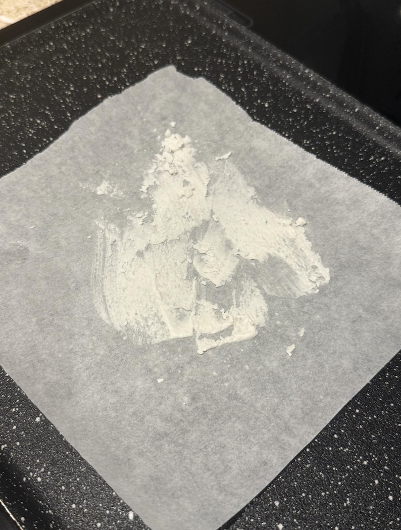 May include: A square of parchment paper with a white, textured substance spread across it. The paper sits on a dark, speckled surface, possibly a baking sheet or tray. The substance appears to be a paste or powder.