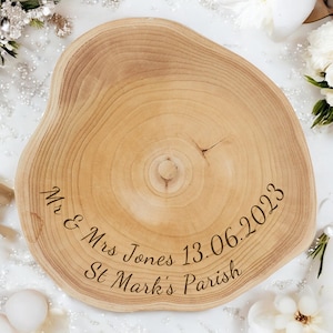 May include: A light brown, wooden, tree-slice serving board with engraved text. The text reads "Mr & Mrs Jones 13.06.2023 St Mark's Parish". The board has a natural, organic shape and visible wood grain.