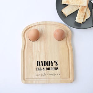 Puede incluir: Bandeja de servir de madera para huevos y soldados con forma de rebanada de pan tostado. La bandeja tiene dos hendiduras para los huevos y el texto "Daddy's Egg & Soldiers Love Alfie & Amelia x x" está grabado en la parte delantera.