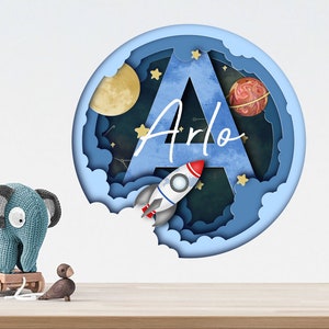 May include: A blue and white paper cut-out wall decal featuring a large letter A with a rocket, stars, and planets. The name "Arlo" is written in cursive on the decal.