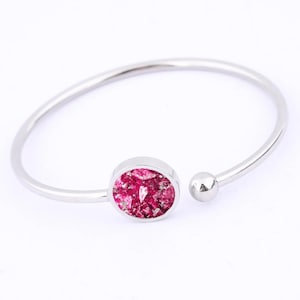 Könnte beinhalten: Ein silberfarbenes, offenes Armreif-Armband mit einer zentralen, ovalen Fassung, die mit einem strukturierten, rosa Material gefüllt ist. Das Armband hat eine glatte, polierte Oberfläche und ein kleines Kugeldetail am offenen Ende. Ein Schmuckstück.