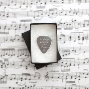 Könnte beinhalten: Ein silberner Gitarrenplektrum mit dem Text "I'd always pick you! Love Emma xx" eingraviert. Das Plektrum befindet sich in einer schwarzen Box auf einem Notenblatt.