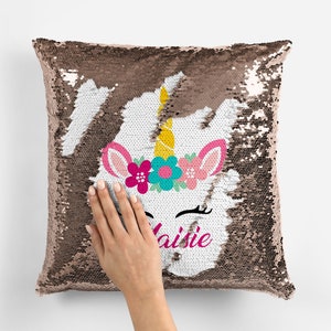 Op de afbeelding: Een kussen van witte en roze gouden sequins met een eenhoorn gezicht en de naam Maisie erop geborduurd. De eenhoorn heeft een roze bloemenkroon en een gele hoorn.