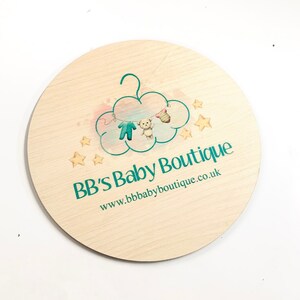 May include: A round wooden sign with a teal blue logo that says "BB's Baby Boutique" and the website address "www.bbbabyboutique.co.uk". The logo features a cloud with a hanger and baby clothes hanging from it.