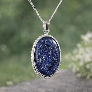 May include: A silver pendant necklace with an oval, dark blue stone with a speckled pattern. The stone is set in a silver frame with a rope-like border and hangs from a delicate silver chain. The background is blurred, with green and white tones.