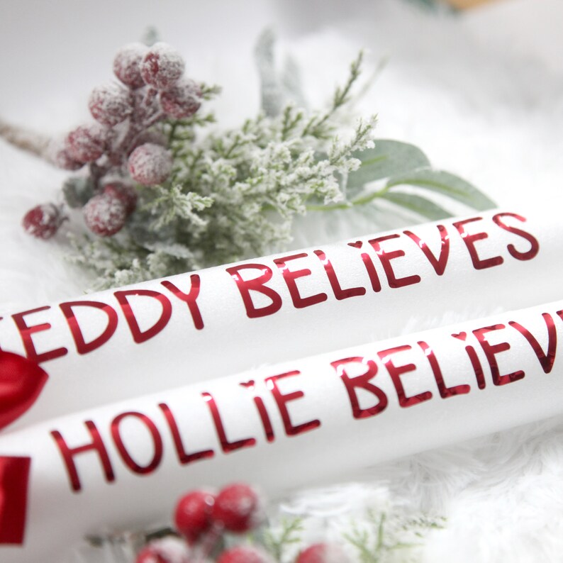 May include: Two white, rolled up paper tubes with red lettering. The top tube says "EDDY BELIEVES" and the bottom tube says "HOLLIE BELIEVES". The tubes are decorated with a sprig of faux greenery with red berries.