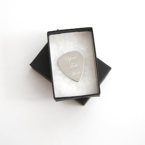 Puede incluir: Una púa de guitarra plateada en una caja de regalo negra. La púa está grabada con el texto "Your Text Here".