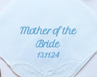 May include: White handkerchief with blue embroidered text reading "Mother of the Bride" and the date "13.11.24". The handkerchief has a delicate white lace border. Ideal for a wedding.