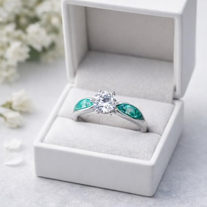 May include: A silver ring with a large, clear central stone and two teal-coloured side stones, displayed in a white ring box. The ring is set against a soft, blurred background.