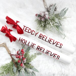 May include: Two white Christmas gift tubes with red bows and the text "Teddy Believes" and "Hollie Believes" in red lettering.