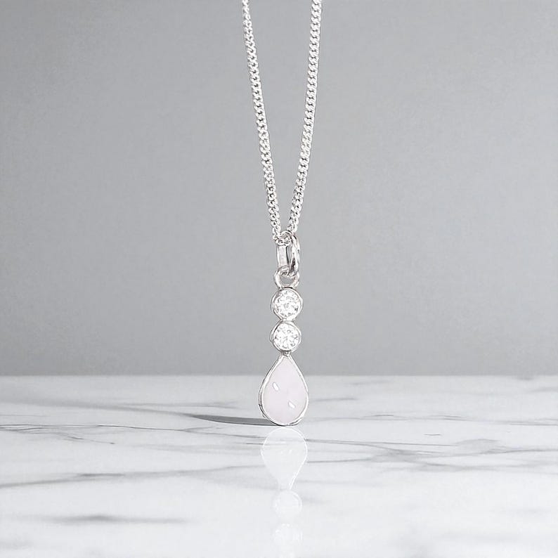 May include: A silver necklace with a pendant featuring two round, clear stones above a pear-shaped, light pink stone. The chain is a delicate curb link style, and the pendant hangs against a light gray background.