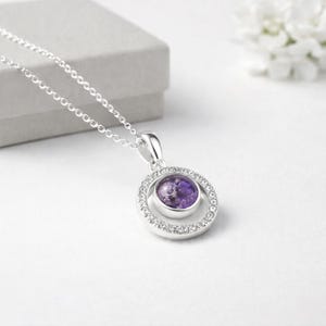 May include: A silver necklace with a circular pendant. The pendant has a central purple stone encircled by small, clear crystals. The chain and pendant are made of shiny silver metal. The necklace is displayed on a white surface.