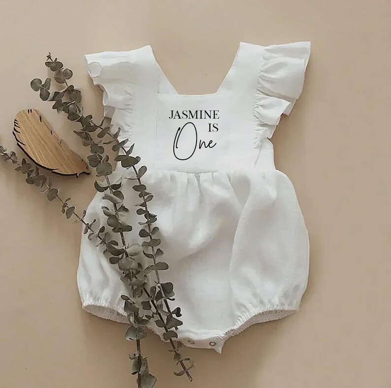 May include: White linen baby onesie with ruffled sleeves and a white patch on the front that says "JASMINE IS One" in black text.