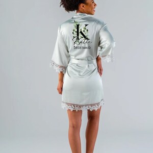 May include: A light green satin robe with a white lace trim. The robe features a floral design with the letter "K" and the name "Katie" written in black. The text "BRIDESMAID" is also written in black.