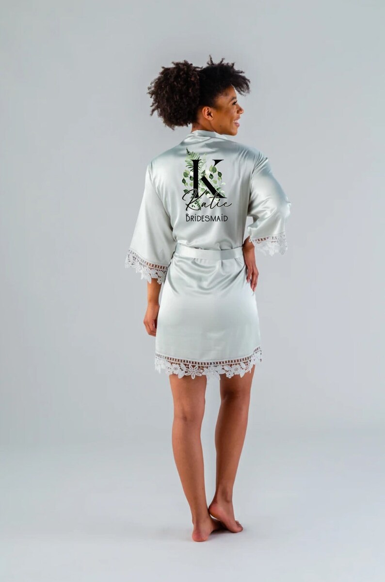 May include: A light green satin robe with a white lace trim. The robe features a floral design with the letter "K" and the name "Katie" written in black. The text "BRIDESMAID" is also written in black.