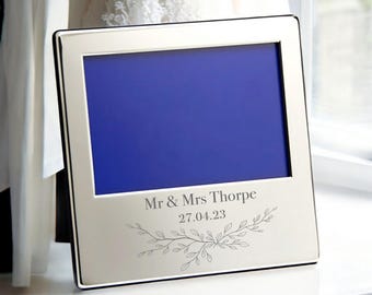 May include: A silver-colored picture frame with a blue interior. The frame has the text "Mr & Mrs Thorpe" and the date "27.04.23" with a decorative floral design. The frame is sitting on a white surface.