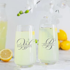 May include: Two clear stemless glasses filled with a yellow liquid, garnished with lemon slices and herbs. The glasses are personalized with a monogram and name: "Ellis Watson" and "Eric Booth". A pitcher and lemons are in the background.