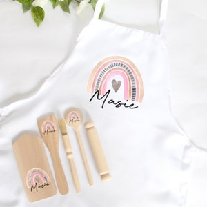 Può includere: Un grembiule bianco con un design a arcobaleno e il nome "Masie" stampato su di esso. Il grembiule è accompagnato da un set di quattro utensili da cucina in legno: un cucchiaio, una spatola, un mattarello e un pennello. Ogni utensile ha un design a arcobaleno e il nome "Masie" stampato su di esso.
