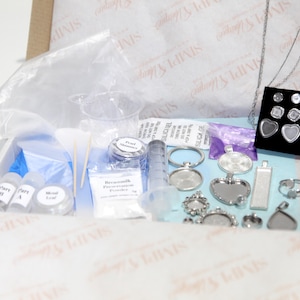 May include: A jewelry-making kit with various components, including silver-toned pendants, rings, and earring settings. The kit also contains small bottles, powders, a syringe, and other crafting supplies, all arranged in a box.