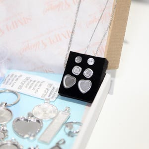 May include: A jewelry making kit with various silver-toned pendants, earrings, and a necklace. The earrings include heart-shaped and square settings. The kit also contains silica gel packets for moisture absorption. The background includes a box and tissue paper.