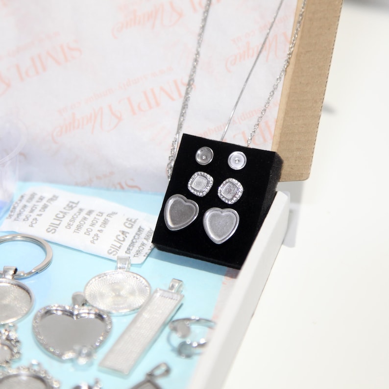 May include: A jewelry making kit with various silver-toned pendants, earrings, and a necklace. The earrings include heart-shaped and square settings. The kit also contains silica gel packets for moisture absorption. The background includes a box and tissue paper.
